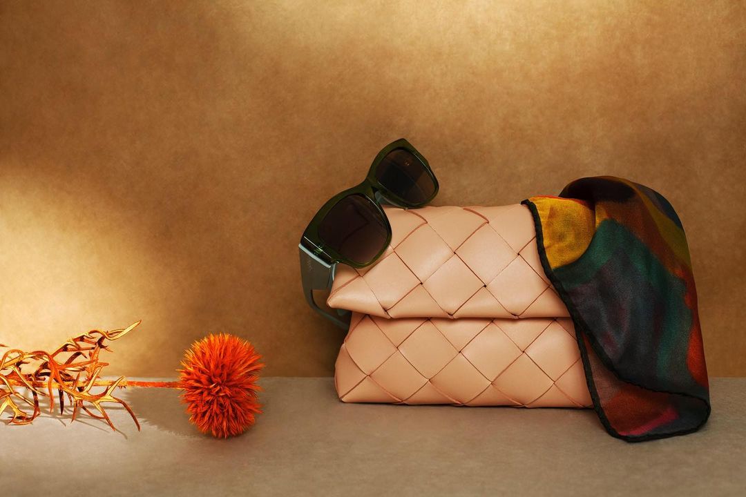 Studio shot featuring a light pink woven handbag, sunglasses, and silk scarf against a tan background, accentuated by dried floral accents.