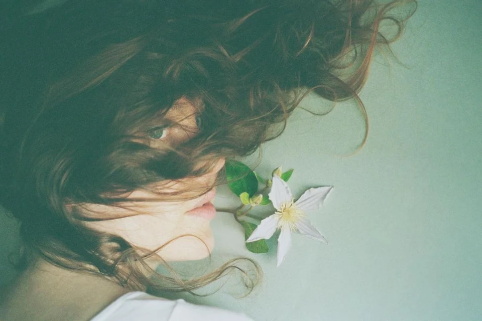 Portrait of a woman with curly hair lying on a light green surface, accented by a white flower near her face in a soft, dreamy aesthetic.