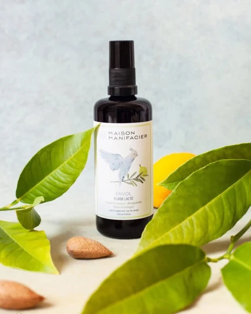 Bottle of Maison Manifacier Envol Fluide Lacte surrounded by green leaves and an almond on a light-colored surface.