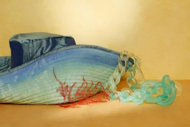 Blue quilted bucket hat with translucent plastic chain and red dried plant against yellow wall