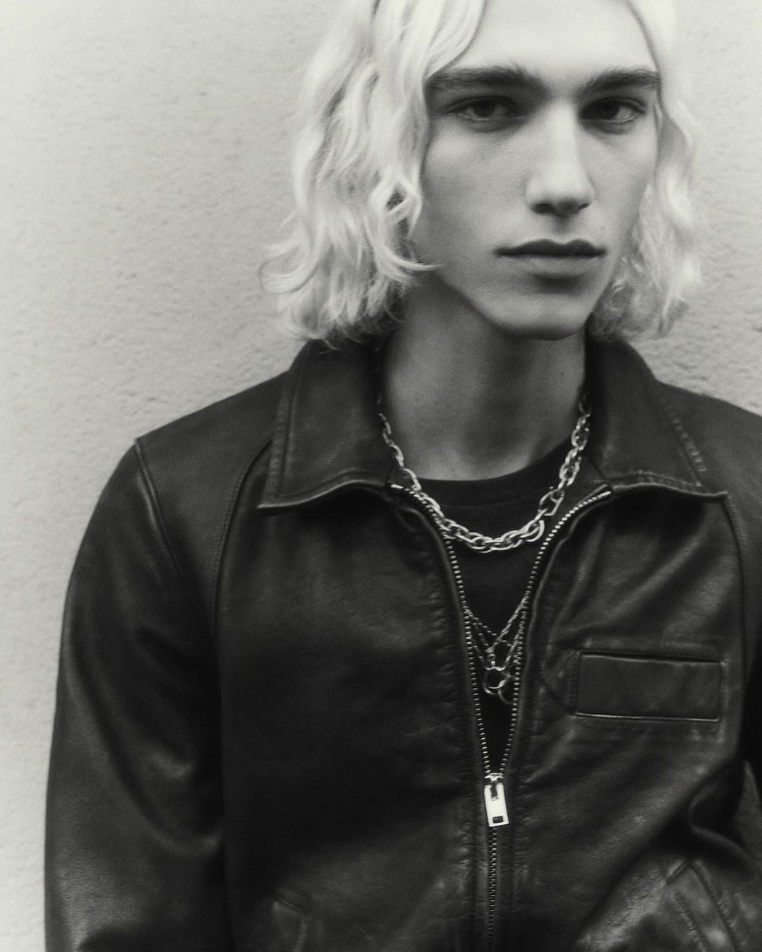 Portrait of a young man with wavy blonde hair wearing a black leather jacket and silver chain necklaces