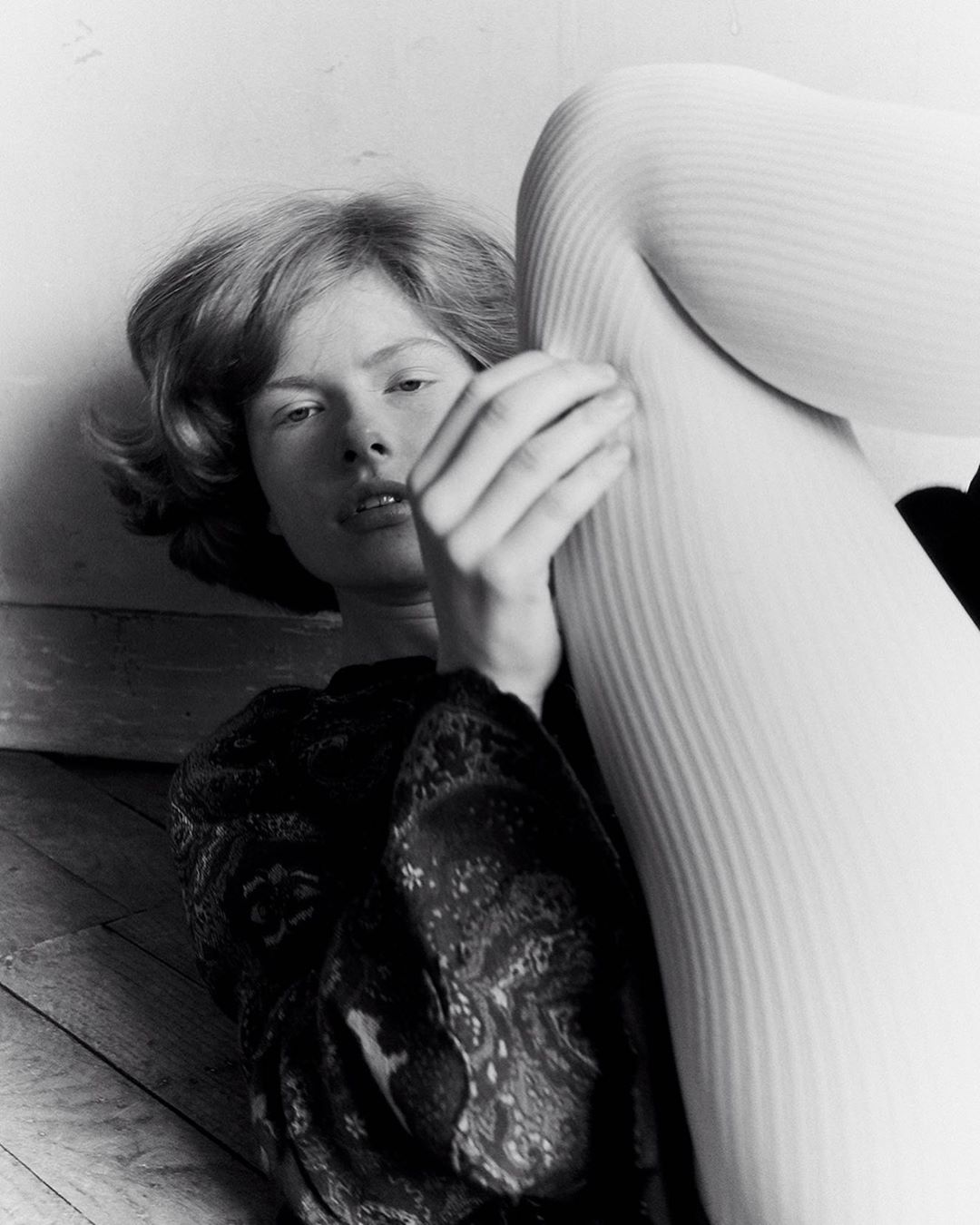 Artistic black and white studio portrait of a young adult woman with wavy hair lying next to a leg clad in white ribbed stockings.