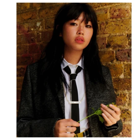 Portrait of a young woman with dark hair wearing a grey blazer, white shirt, and black tie against a brick wall background.