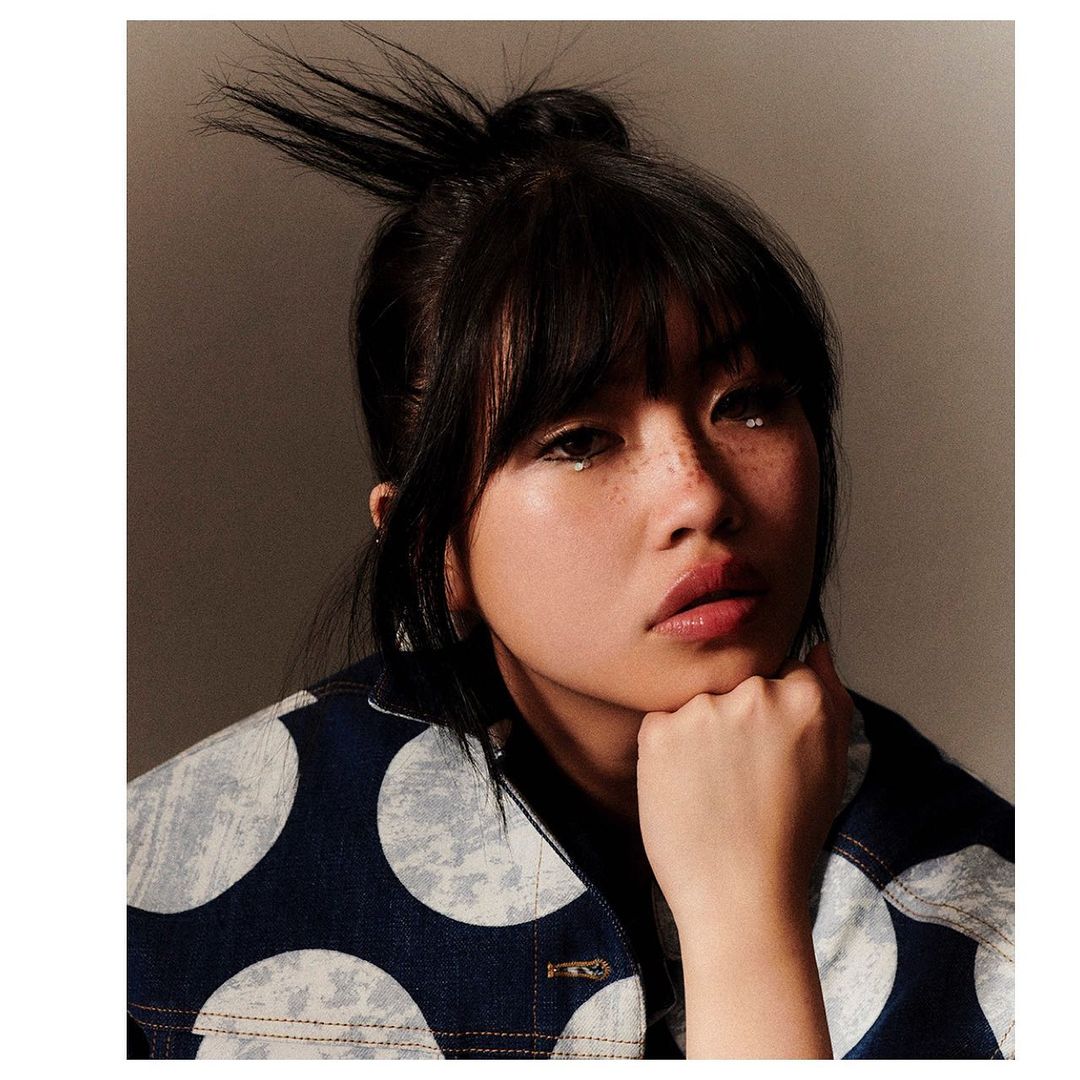 Close-up portrait of a young woman with dark hair and bangs, wearing a blue and white polka dot jacket, against a neutral backdrop.