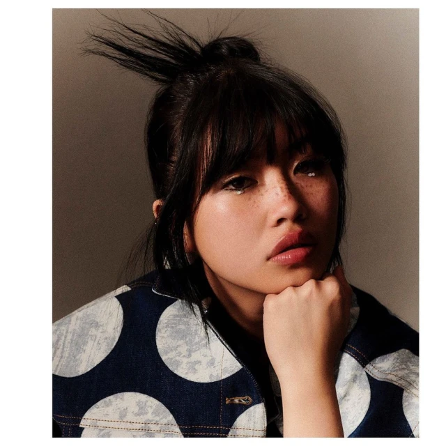 Close-up portrait of a young woman with dark hair and bangs, wearing a blue and white polka dot jacket, against a neutral backdrop.