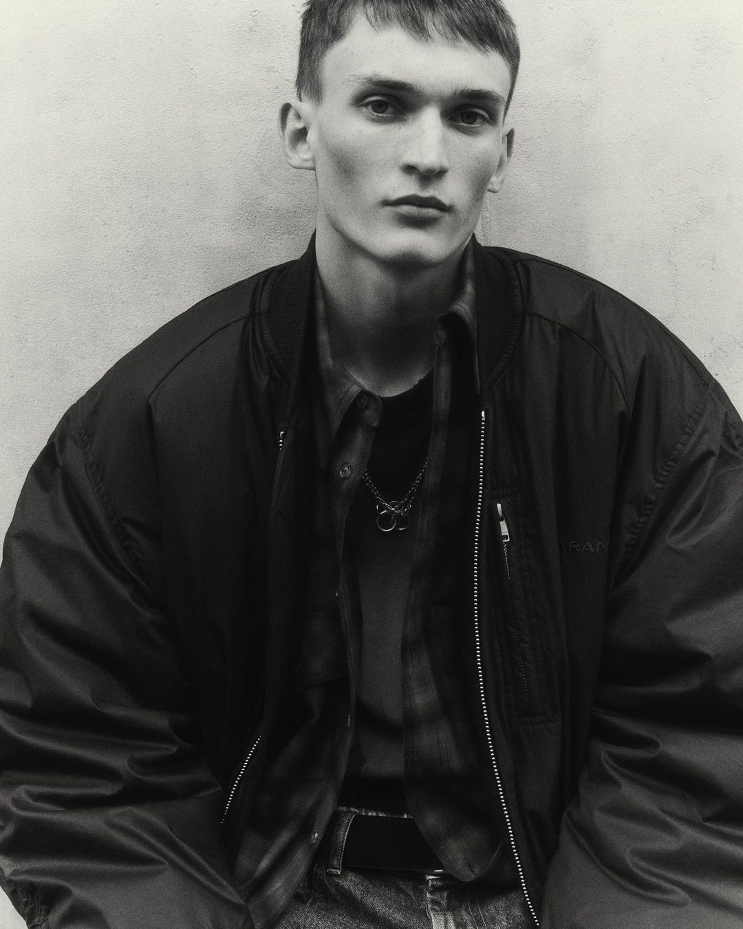 Monochrome portrait of a young man wearing a dark zip-up bomber jacket and a layered collared shirt against a plain textured wall