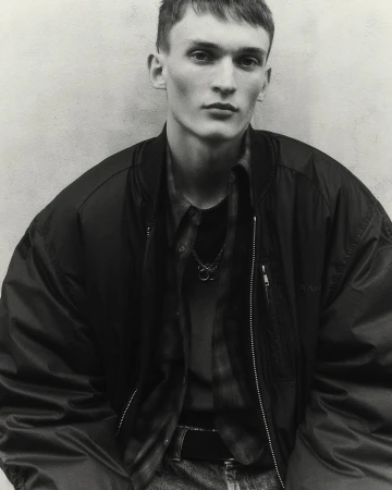 Monochrome portrait of a young man wearing a dark zip-up bomber jacket and a layered collared shirt against a plain textured wall