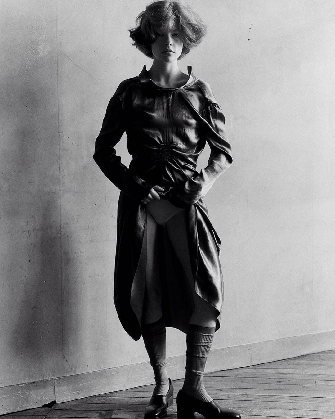Full-length portrait of a young woman in a dress, knee socks, and platform shoes. The black and white photo has soft, diffused lighting.