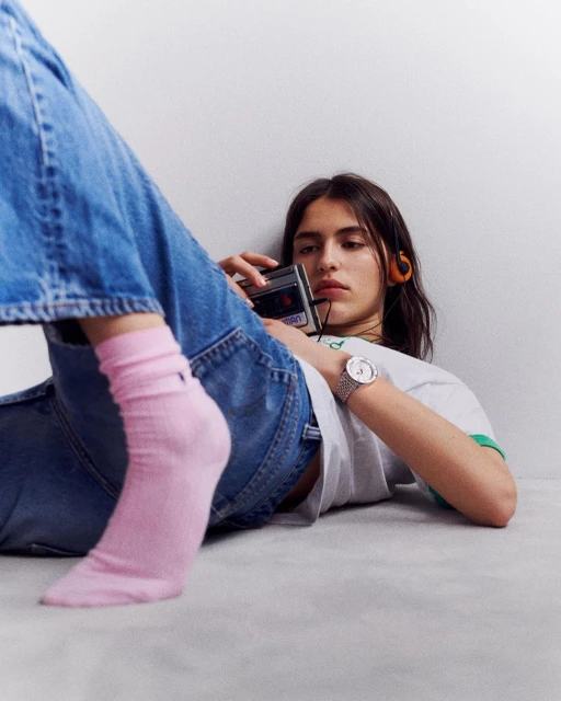 Brunette woman lying on her side on a gray floor wearing pink socks and jeans, holding a vintage cassette player with headphones. 