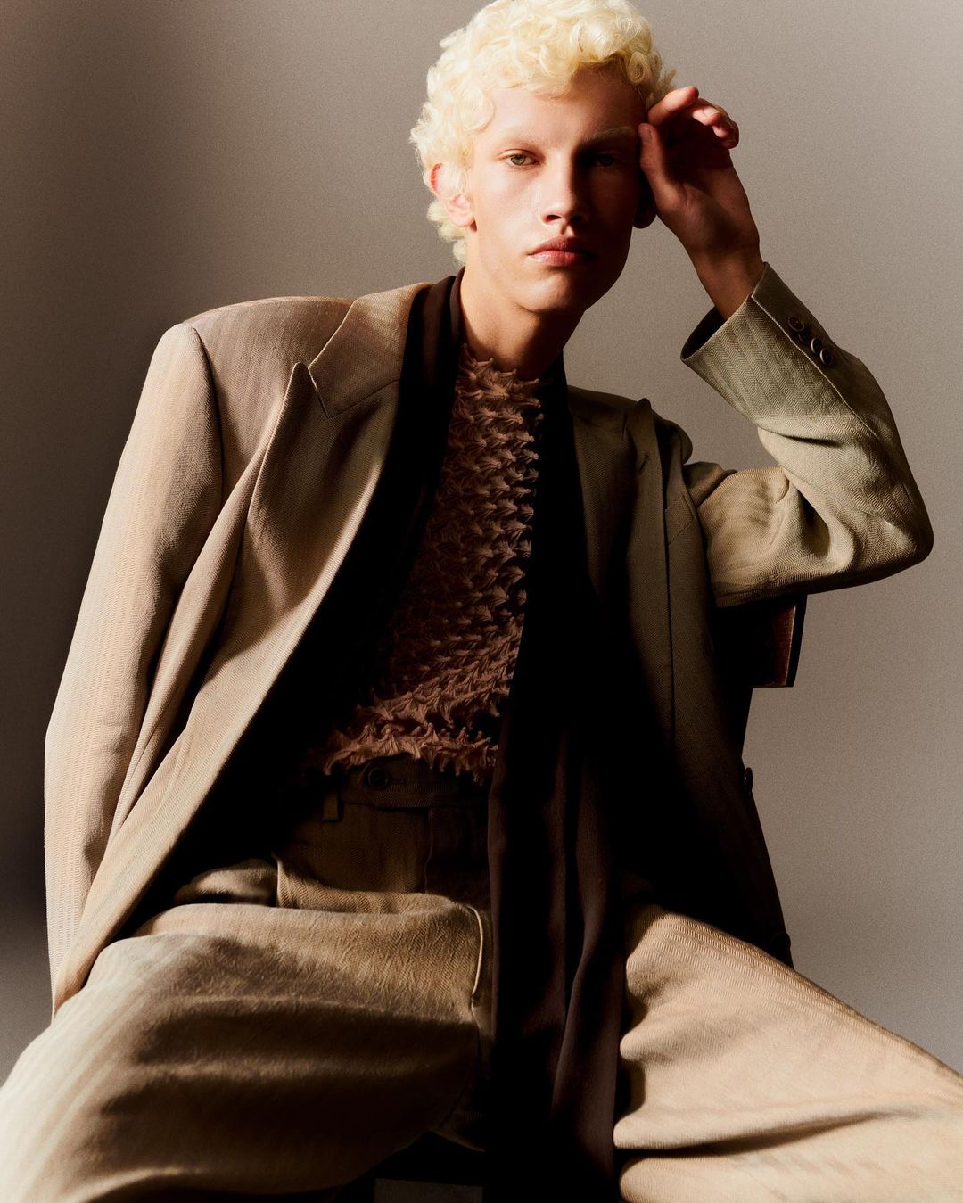 Young adult man in stylish neutral-toned suit posing indoors, hand on head, with light blond curly hair. Fashion editorial style.