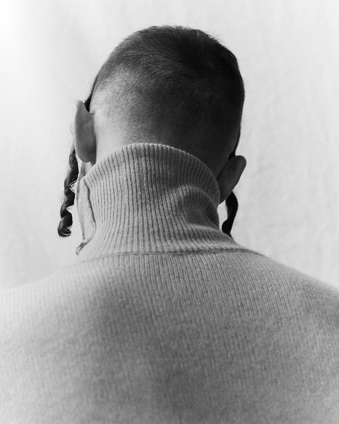 Back view of an adult with buzz-cut hair, visible sidecurls, and wearing a tan turtleneck sweater against a plain white backdrop.