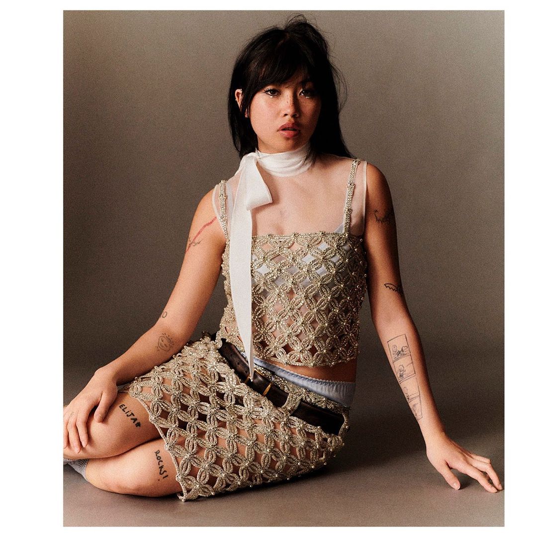 Portrait of an Asian woman with dark hair, wearing a beaded top and skirt with arm tattoos, posing in a photo studio.