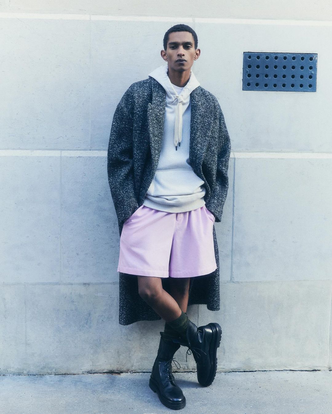 Dark-skinned adult man posing in an overcoat, hoodie, and shorts against a textured light-grey wall in natural light. Fashion editorial.