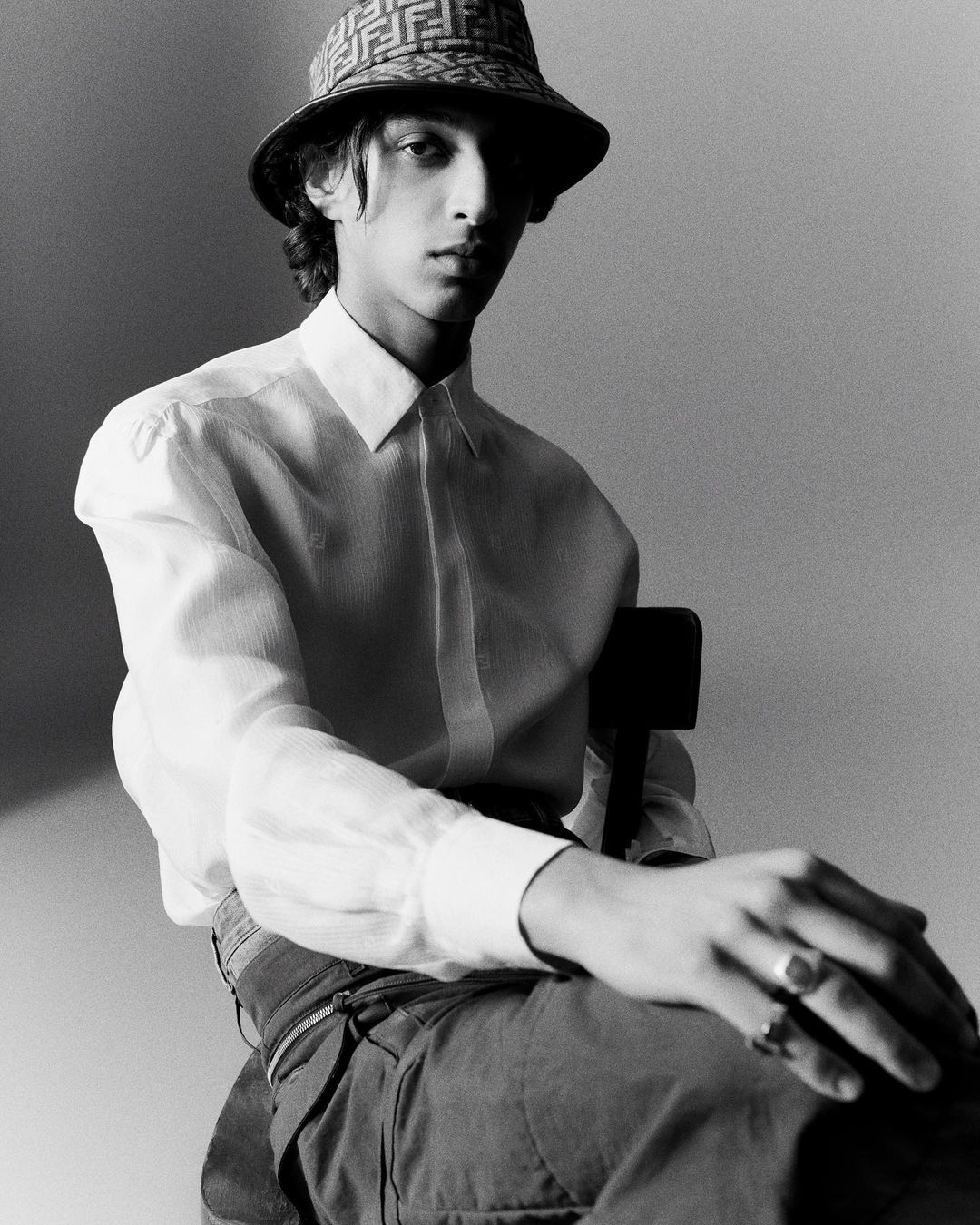 Young man in a white shirt and patterned hat sits in a minimalist gray studio, looking directly at the camera in a fashion-style portrait.