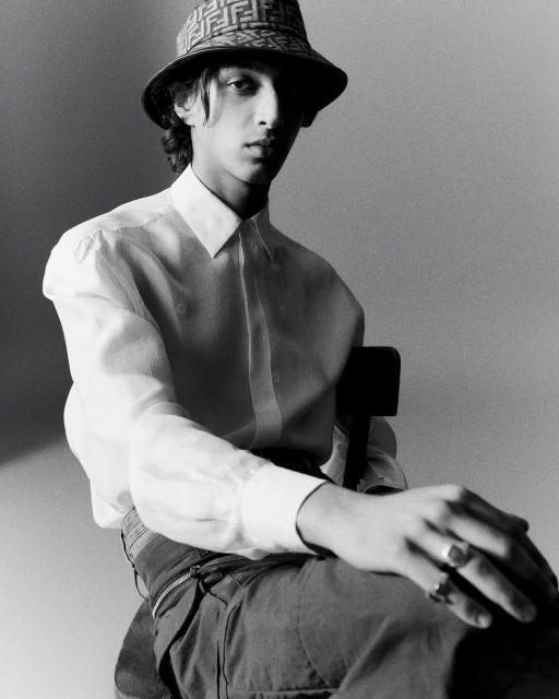 Young man in a white shirt and patterned hat sits in a minimalist gray studio, looking directly at the camera in a fashion-style portrait.