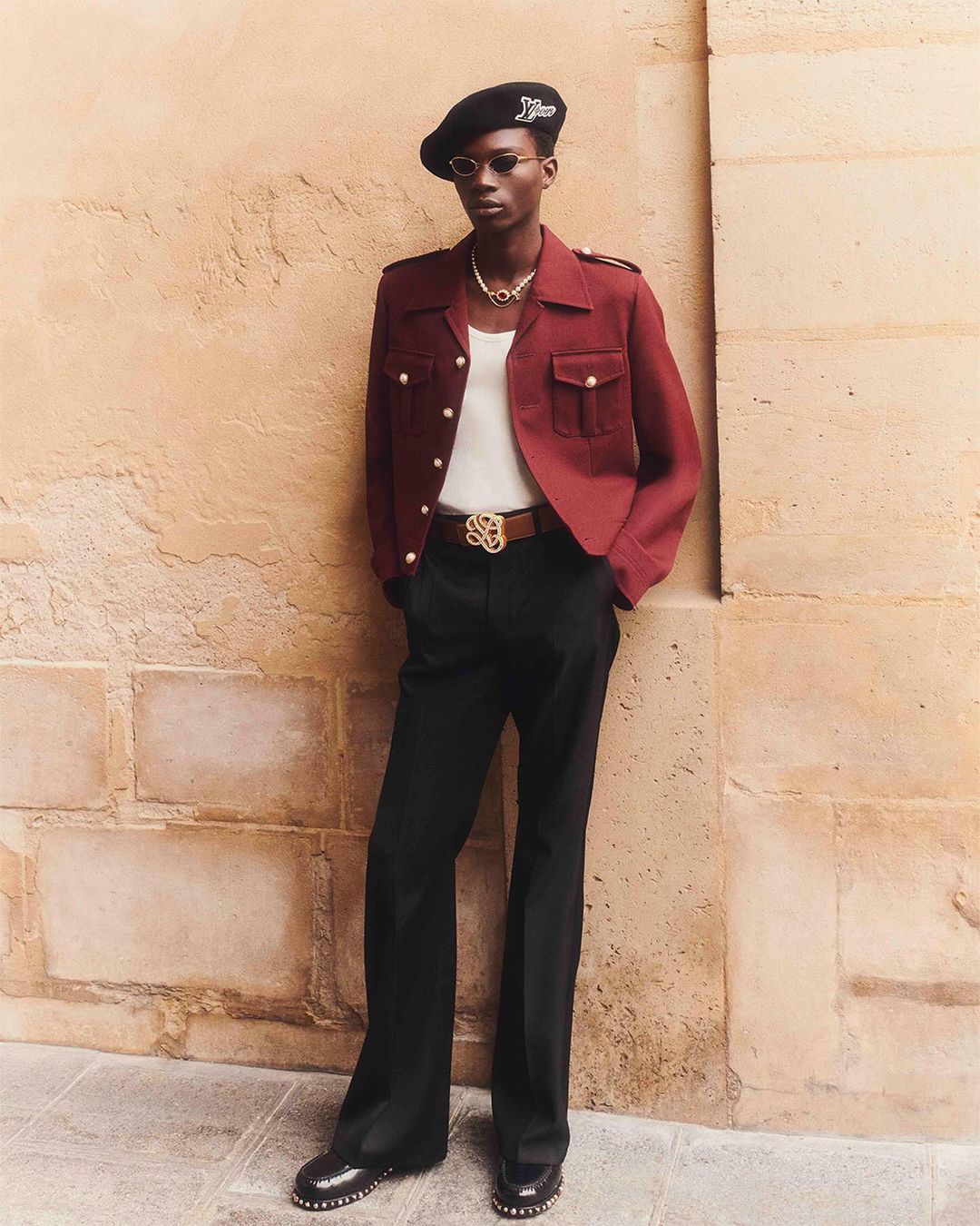 A stylish Black man in a beret, burgundy jacket, black pants, and gold jewelry poses against a tan brick wall in an outdoor setting.