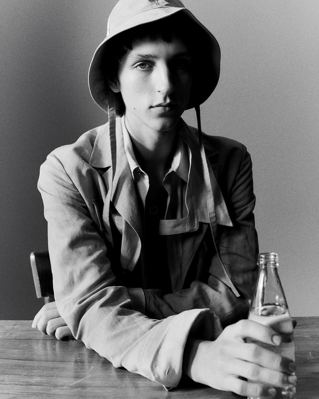 Monochrome portrait of a young man wearing a bucket hat and jacket, seated at a wooden table holding a glass bottle in Paris.