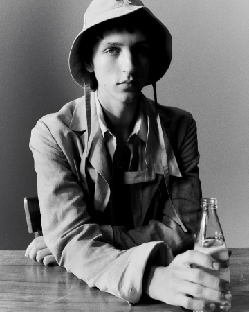Monochrome portrait of a young man wearing a bucket hat and jacket, seated at a wooden table holding a glass bottle in Paris.