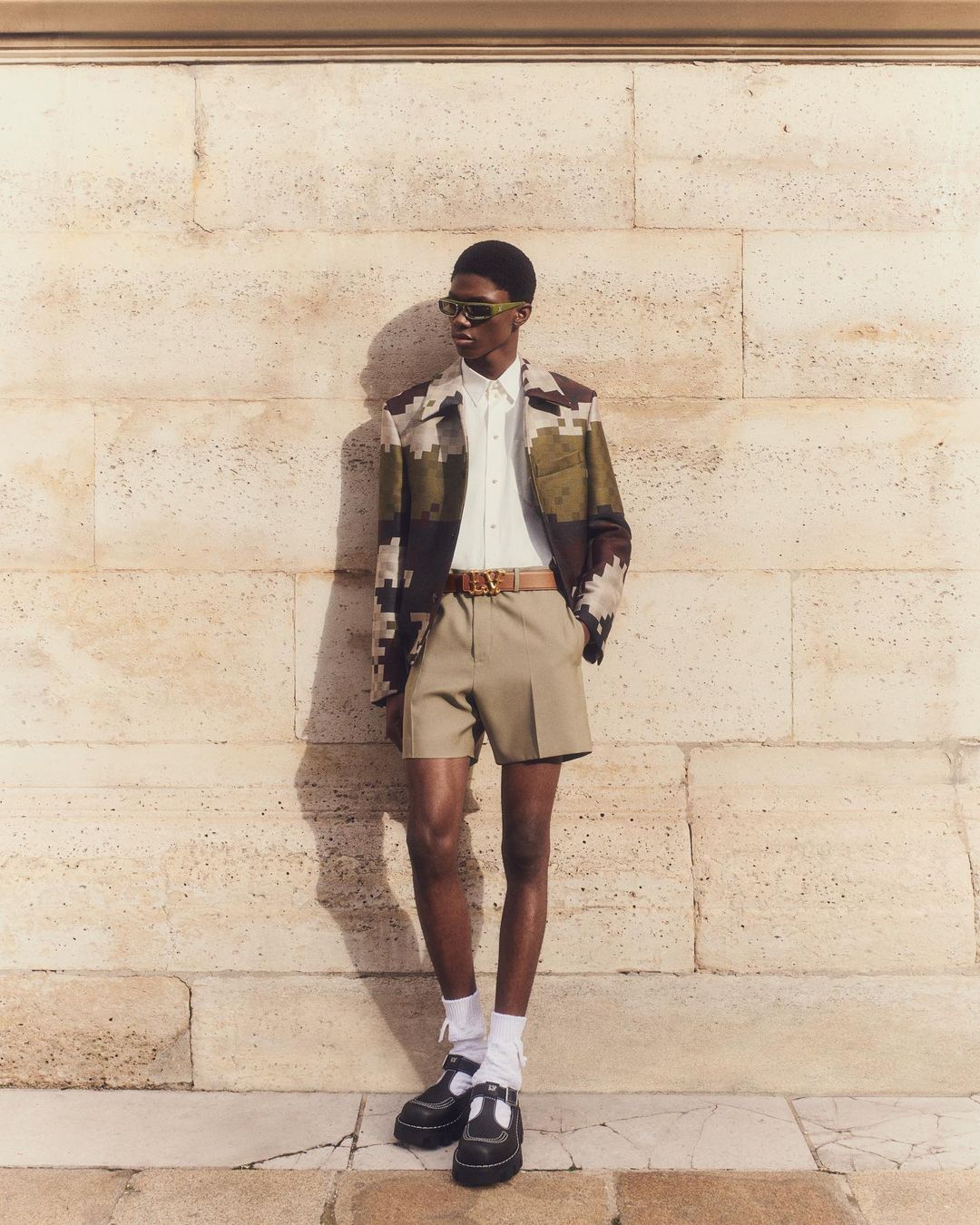 A fashionable adult man stands in front of a stone wall wearing sunglasses, a patterned jacket, shorts, socks, and black Mary Jane shoes.