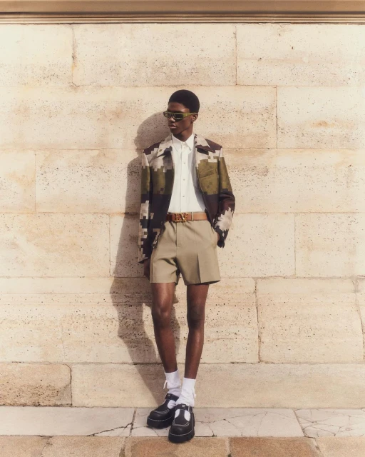 A fashionable adult man stands in front of a stone wall wearing sunglasses, a patterned jacket, shorts, socks, and black Mary Jane shoes.