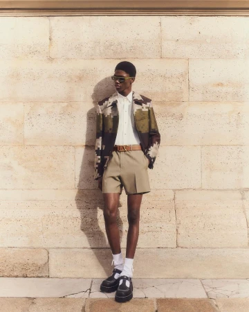 A fashionable adult man stands in front of a stone wall wearing sunglasses, a patterned jacket, shorts, socks, and black Mary Jane shoes.