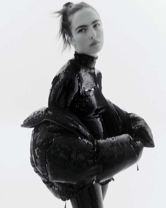 Monochrome portrait of a female model in a shiny, black, textured, and unique puffer jacket against a bright, neutral backdrop.
