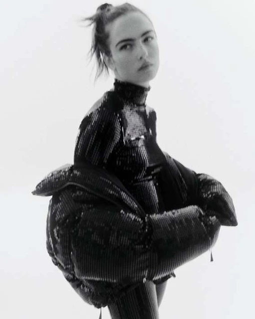 Monochrome portrait of a female model in a shiny, black, textured, and unique puffer jacket against a bright, neutral backdrop.