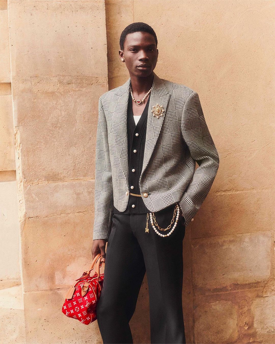 Fashionable Black man poses against a neutral wall in a patterned blazer, vest, and red handbag, showcasing style and elegance.