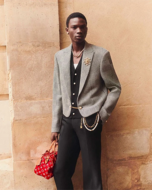 Fashionable Black man poses against a neutral wall in a patterned blazer, vest, and red handbag, showcasing style and elegance.