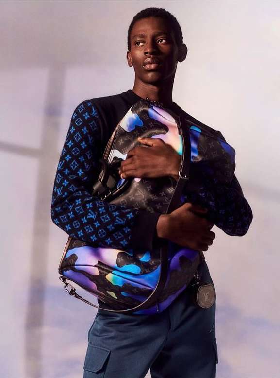 Man poses with a patterned duffel bag in a studio. He's wearing a designer sweater and dark pants, fashion photography, studio lighting.