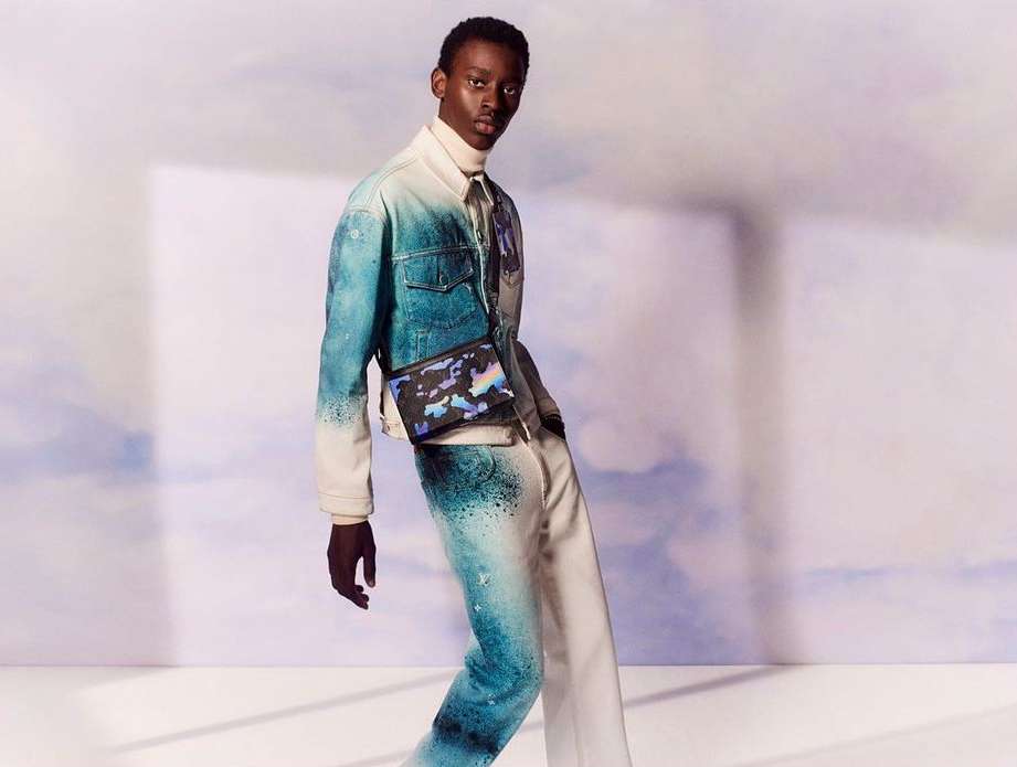 Male fashion model poses in a blue and white ombre dyed denim jacket and pants with a crossbody bag against a gradient backdrop.
