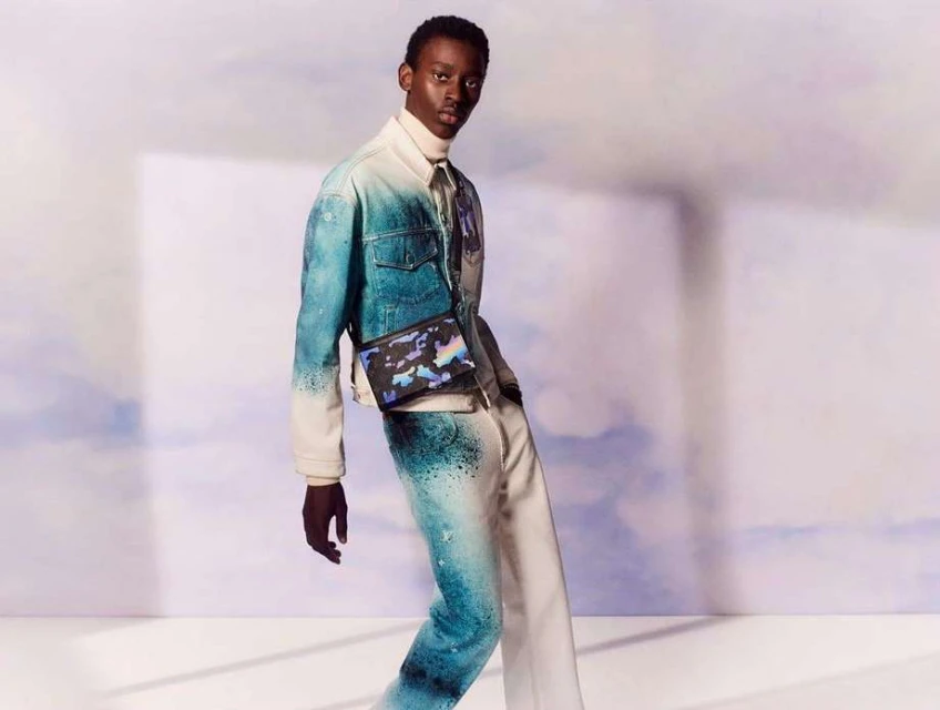 Male fashion model poses in a blue and white ombre dyed denim jacket and pants with a crossbody bag against a gradient backdrop.