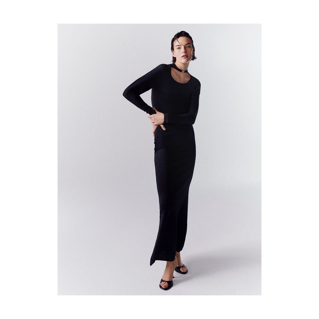 A young adult woman poses in a long-sleeved, floor-length black dress with a white studio background, her hand resting on her hip.