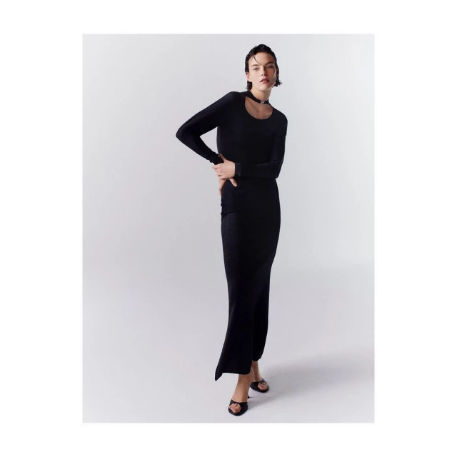 A young adult woman poses in a long-sleeved, floor-length black dress with a white studio background, her hand resting on her hip.