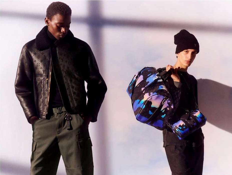 Two fashion models pose in a studio illuminated by directed lighting. One carries patterned duffel bags; the other wears a logo jacket.