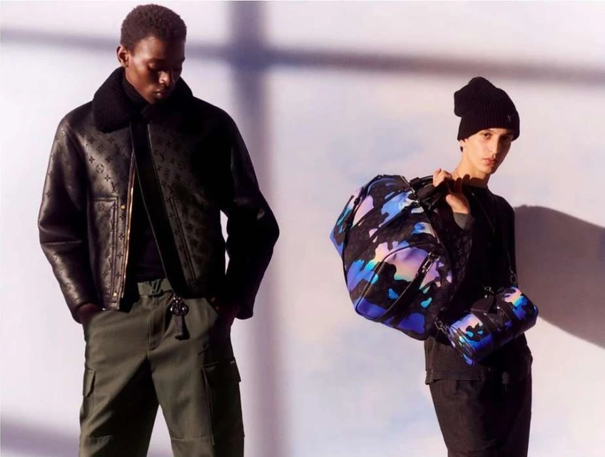 Two fashion models pose in a studio illuminated by directed lighting. One carries patterned duffel bags; the other wears a logo jacket.