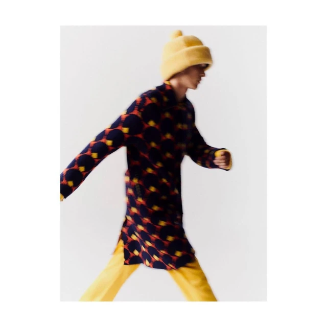 Fashionable adult walks in a patterned tunic and matching beanie hat, with yellow pants against a clean white backdrop.