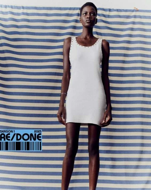 Fashion shot of Black female model in a white mini dress against a striped blue and white background. Re/Done logo with barcode visible.