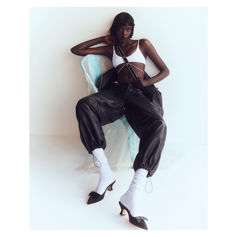 Fashion shot of Black female model in white top, oversized leather pants, socks, and black heels, sitting indoors with arm raised.