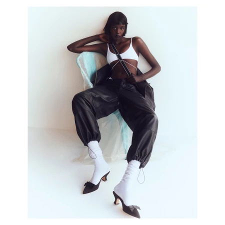 Fashion shot of Black female model in white top, oversized leather pants, socks, and black heels, sitting indoors with arm raised.