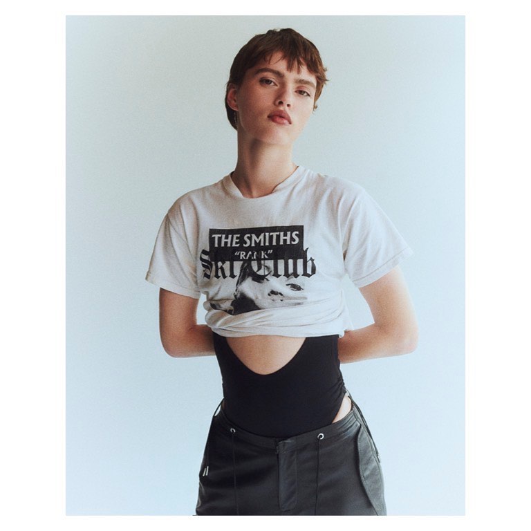 Model with short brown hair wearing a graphic white t-shirt and black pants against a seamless white background.