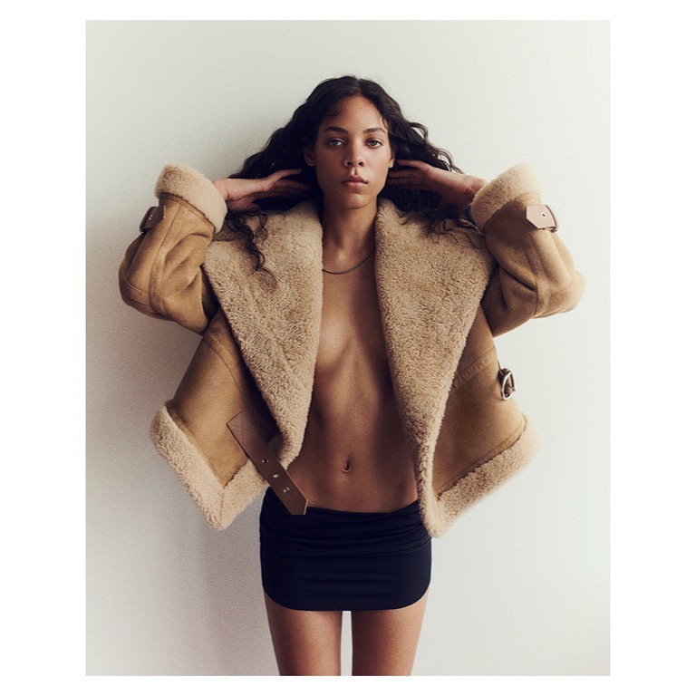 Woman in shearling jacket and black mini-skirt poses against a white background with her hands behind her head.