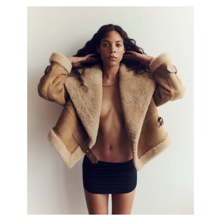 Woman in shearling jacket and black mini-skirt poses against a white background with her hands behind her head.