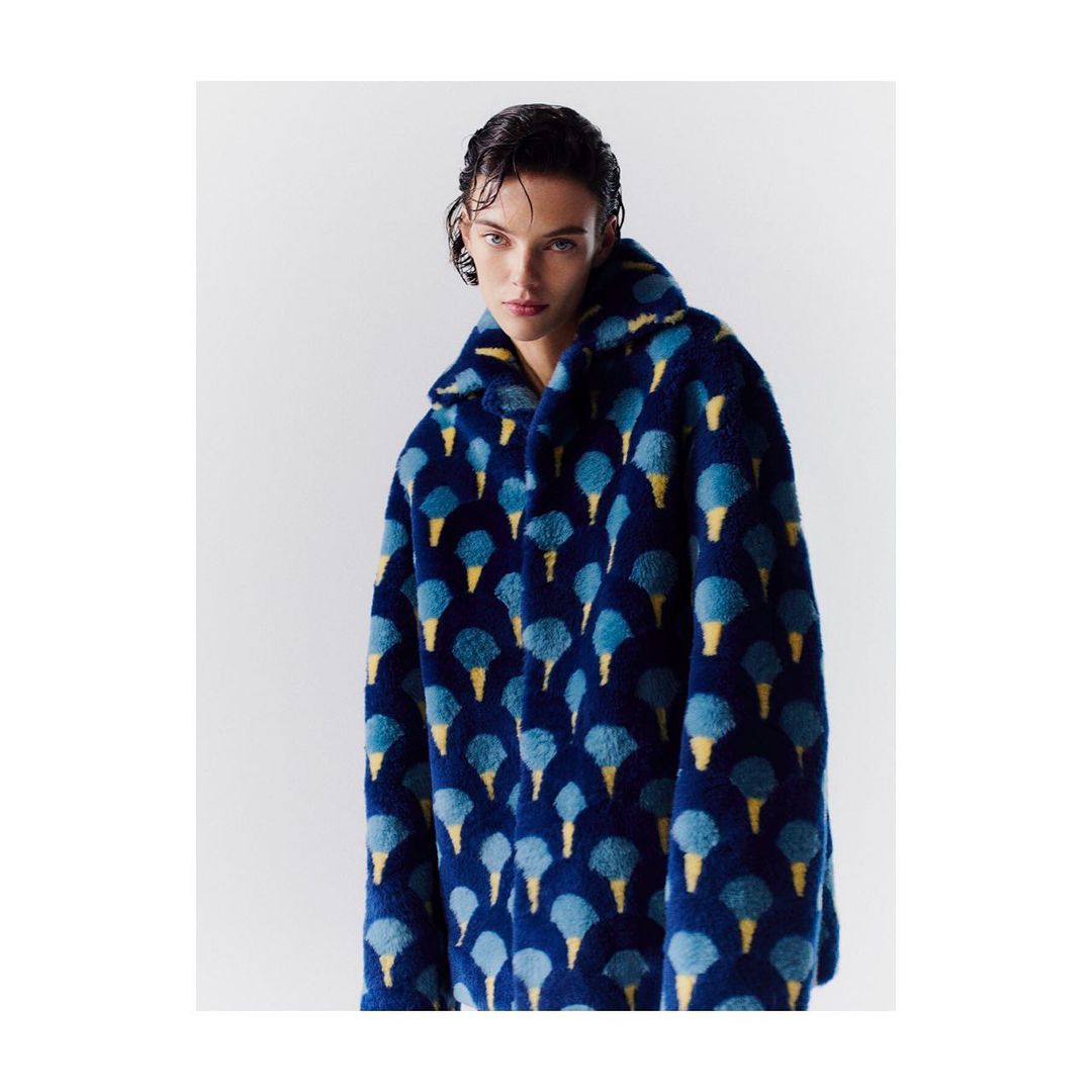 Woman poses in a studio wearing a blue faux fur coat with an ice cream cone pattern. Neutral background.