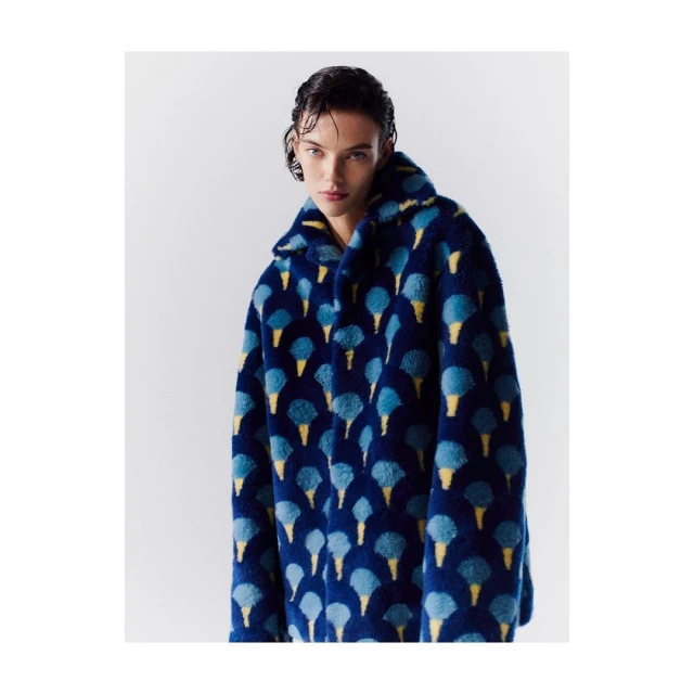 Woman poses in a studio wearing a blue faux fur coat with an ice cream cone pattern.  Neutral background.