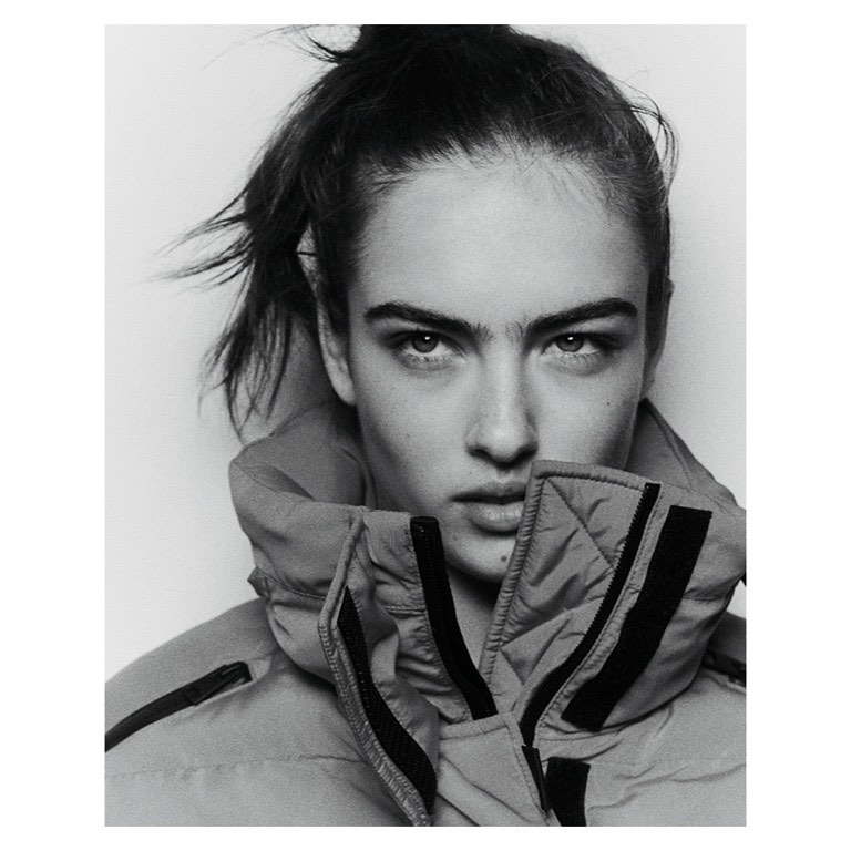 Black and white portrait of a young woman with her hair up, wearing a high collared puffer jacket against a white backdrop.