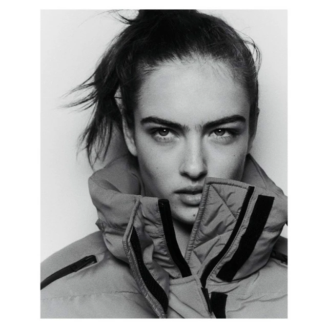Black and white portrait of a young woman with her hair up, wearing a high collared puffer jacket against a white backdrop.
