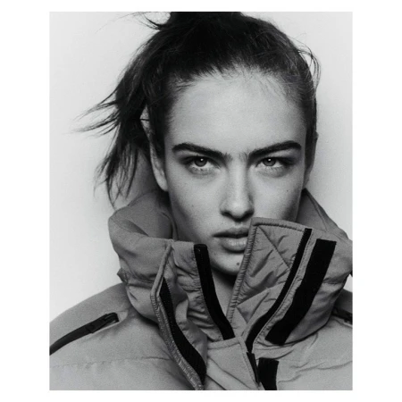 Black and white portrait of a young woman with her hair up, wearing a high collared puffer jacket against a white backdrop.