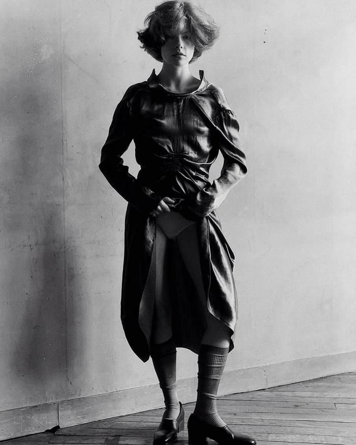 Monochrome fashion portrait of a model in a dark dress and knee-high socks standing against a textured wall