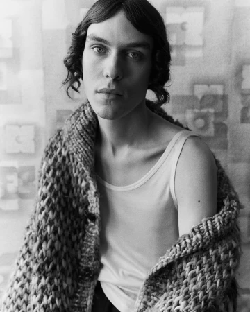 Black and white portrait of a man wearing a white tank top and a heavy knitted cardigan draped over his shoulders.